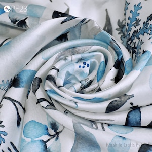 Puede incluir: Tela de seda blanca con estampado floral de acuarela azul y gris. La tela está doblada y drapeada, mostrando la textura y el patrón.