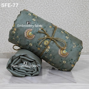 May include: Two rolls of fabric, one embroidered and one plain, in a muted teal color. The embroidered fabric is adorned with floral patterns in yellow, pink, and green. The rolls are tied with twine, and the text "Embroidery fabric" and "Plain matching fabric" are visible.
