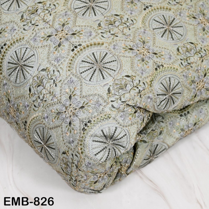 May include: Close-up of a folded fabric with an intricate embroidered design. The fabric features a pale green background with a repeating pattern of floral and geometric shapes in silver, black, and gold thread. The text "EMB-826" is visible.