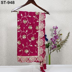 Puede incluir: Un textil magenta con intrincados bordados florales dorados y un borde festoneado y adornado. La tela est&aacute; drapeada sobre una percha de madera. El texto "ST-948" es visible en la esquina superior izquierda. Un ramo de flores est&aacute; en el fondo.