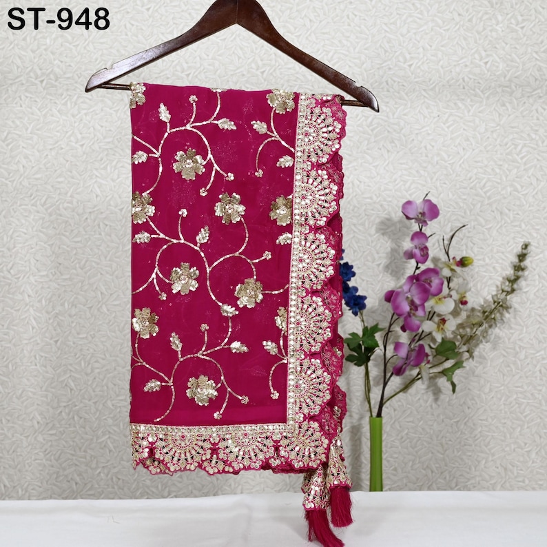 Puede incluir: Un textil magenta con intrincados bordados florales dorados y un borde festoneado y adornado. La tela est&aacute; drapeada sobre una percha de madera. El texto "ST-948" es visible en la esquina superior izquierda. Un ramo de flores est&aacute; en el fondo.