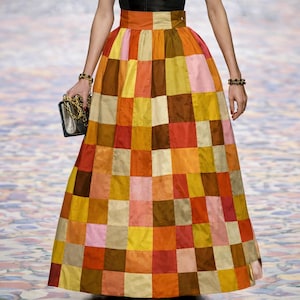 May include: A long, patchwork skirt in a variety of warm colors, including orange, yellow, brown, and pink. The skirt is made of squares of fabric and has a high waist. A small, black purse with a gold chain is held in the left hand.