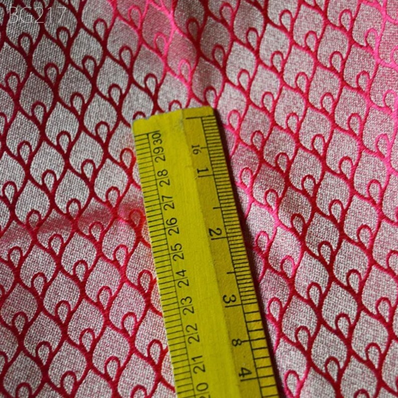 May include: Close-up of a pink fabric with a repeating pattern of red hearts. A yellow ruler is placed on the fabric, showing measurements in inches.