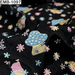 May include: Black fabric featuring an embroidered pattern of light blue mushrooms with white dots and small pink, yellow, and white flowers. The text EMB-1097 is visible in the top left corner. The material has a textured surface with folds.
