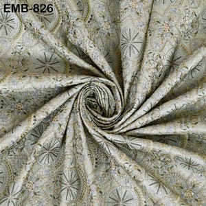 May include: Close-up of a fabric with an intricate embroidered design. The fabric is a light green color with a pattern of circular and floral motifs. The embroidery includes silver, gold, and green threads, creating a textured and detailed appearance. The text "EMB-826" is visible.