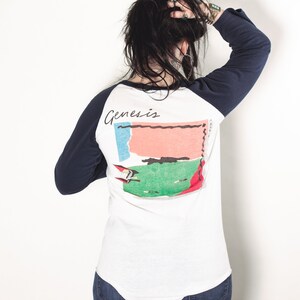 May include: A white baseball tee shirt with navy blue sleeves. The back of the shirt features a graphic of a colorful abstract design with the word "Genesis" printed above it.