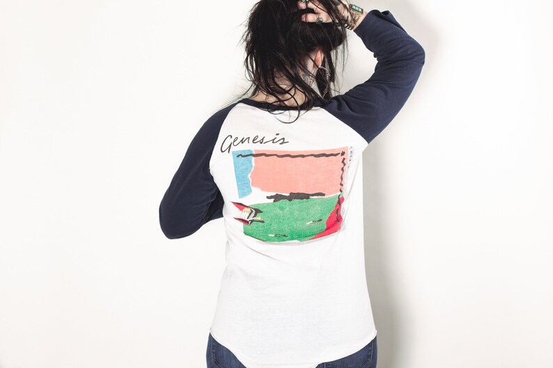 May include: A white baseball tee shirt with navy blue sleeves. The back of the shirt features a graphic of a colorful abstract design with the word "Genesis" printed above it.