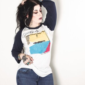 May include: A woman wearing a white and blue baseball tee shirt with the text "Genes is" and a graphic of a map. She is wearing blue jeans and has long black hair.