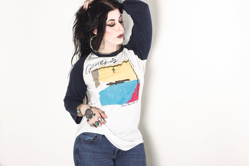May include: A woman wearing a white and blue baseball tee shirt with the text "Genes is" and a graphic of a map. She is wearing blue jeans and has long black hair.