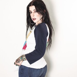 May include: A woman with long black hair wearing a white and blue baseball shirt with a graphic print and blue jeans. She has a tattoo on her left hand.