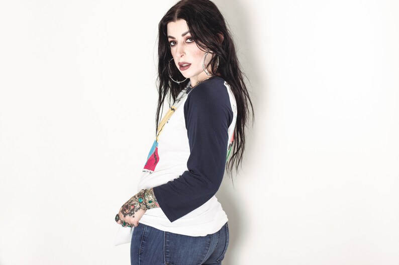 May include: A woman with long black hair wearing a white and blue baseball shirt with a graphic print and blue jeans. She has a tattoo on her left hand.