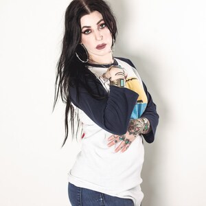 May include: A woman with long black hair wearing a white and blue baseball tee shirt with a graphic design. She is wearing blue jeans and has several rings on her fingers.