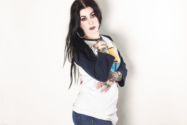 May include: A woman with long black hair wearing a white and blue baseball tee shirt with a graphic design. She is wearing blue jeans and has several rings on her fingers.
