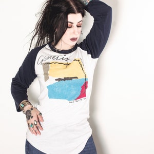 May include: A white and navy blue baseball tee shirt with a colorful graphic design and the text "Genesis - World Tour 1981-82".