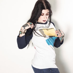 May include: A woman with long black hair wearing a white and blue baseball tee shirt with the word "Genesis" written on it. She is wearing a watch on her left wrist and several rings on her fingers.