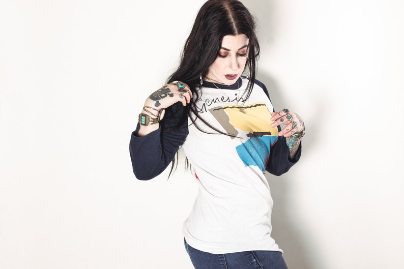 May include: A woman with long black hair wearing a white and blue baseball tee shirt with the word "Genesis" written on it. She is wearing a watch on her left wrist and several rings on her fingers.