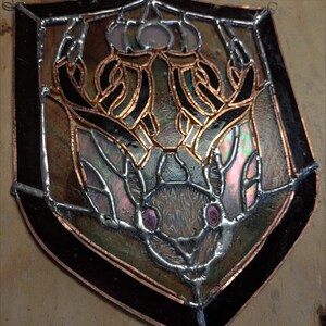 Puede incluir: Una ventana de vitral que representa un murciélago con alas iridiscentes y ojos rosados, rodeado de un marco de cobre y negro.