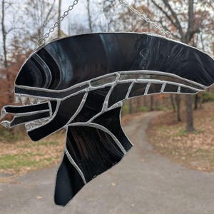 May include: A black stained glass hanging of an alien creature with a detailed face and head. The creature has a large mouth with sharp teeth and a long, thin neck.