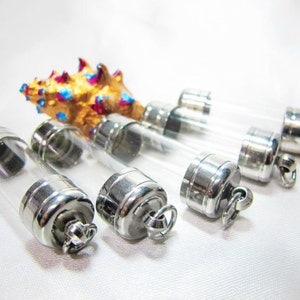 May include: A collection of clear glass vials with silver-colored metal caps and small loops. The vials are cylindrical and appear empty. A decorative seashell with gold, blue, and pink accents is in the background.