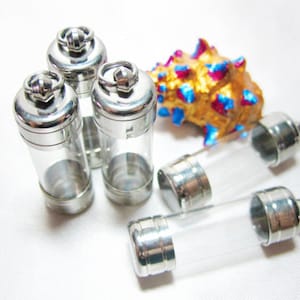 May include: Several small, clear glass vials with silver-colored metal caps and a loop at the top. The vials are cylindrical and appear empty. A decorative gold-colored seashell with blue and pink accents is also present.