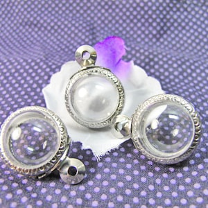 May include: Three silver-toned pendant charms with clear glass domes. The domes are round and have a decorative border. The charms have a loop at the top for attaching to a necklace or bracelet.