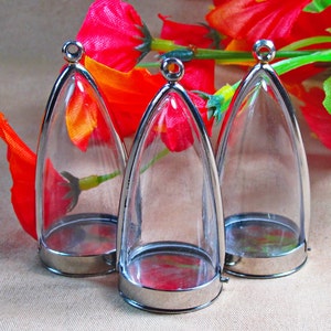 Puede incluir: Tres colgantes de plata con contenedores de vidrio transparente en forma de lágrima. Los contenedores están abiertos en la parte superior y tienen un borde de metal plateado.