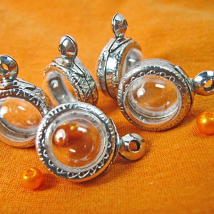 May include: Five silver locket pendants with clear glass inserts. The lockets have a decorative design around the glass.  There are three orange beads on the orange surface.