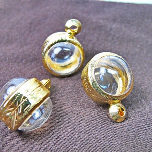 May include: Three gold pendants with clear, spherical glass centres. The gold metal has intricate etched designs. Each pendant has a loop at the top for hanging. The pendants are displayed on a brown fabric surface.