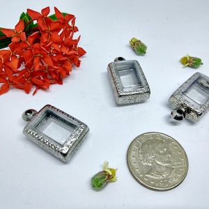 Puede incluir: Tres colgantes plateados con inserciones de vidrio transparente. Los colgantes son rectangulares y tienen un diseño adornado. Los colgantes están sobre una superficie blanca.