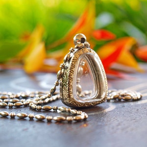 May include: A silver pendant with a clear glass insert, hanging from a silver chain. The pendant has intricate designs and is set against a blurred background of orange and green flowers.