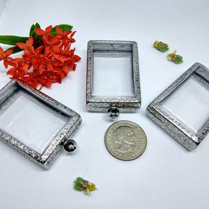 May include: Three silver-toned pendant frames with ornate designs and clear glass inserts. The frames are approximately 2 inches in length and 1.5 inches in width. A US quarter is included for size reference.