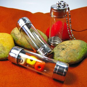 May include: Three silver and glass vials with different contents. The vial on the left contains colorful beads, the middle vial is empty, and the vial on the right contains a red heart. The vials are on a brown fabric surface with two light brown rocks.