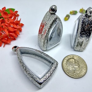 May include: Three silver pendants with clear windows and decorative, patterned edges. Each pendant has a small loop at the top. A coin is next to the pendants. Bright orange flowers are in the background. The pendants are likely for jewellery or display.