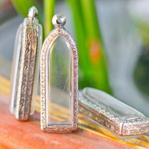 May include: Three silver and glass pendants with intricate designs. The pendants are rectangular and have a clear glass center. The pendants are on a wooden surface.