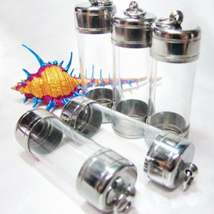 May include: Clear glass vials with silver-colored metal caps and bases. The vials are cylindrical and have a loop at the top of the cap. A decorative seashell with gold, pink, and blue accents is also present.