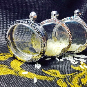 May include: Three silver round pendants with clear glass inserts, filled with white rice grains. The pendants are on a black fabric background.