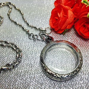 Op de afbeelding: Een zilveren ketting met een hanger in de vorm van een medaillon en een transparante glazen inzet. De ketting is aan een zilveren ketting met ovale schakels bevestigd. Het medaillon is omgeven door een decoratieve rand. De ketting ligt op een zilveren achtergrond.