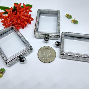 May include: Three silver-toned, rectangular, ornate frames with clear glass inserts. The frames have a decorative border and a small loop at the top. A silver-toned coin is in the center of the image.