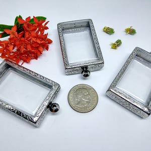 May include: Three rectangular, silver-toned metal lockets with clear inserts, featuring ornate detailing around the edges. A US quarter coin is next to the lockets. Bright orange flowers and small yellow buds are also in the image.