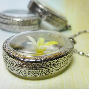 Könnte beinhalten: Eine silberne Halskette mit einem filigranen Blumenmuster. Das Medaillon ist geöffnet und enthält eine gepresste gelbe Blume.