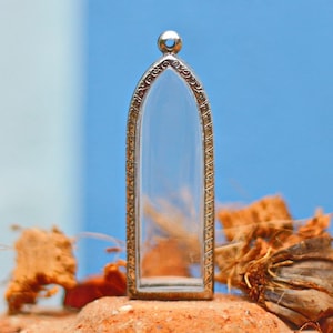 Peut inclure: Un pendentif clair en forme d'arche avec un cadre décoratif de couleur or. Le pendentif a une petite boucle en haut et est vide à l'intérieur. Le fond est un dégradé de bleu et de blanc, avec de la matière organique brune en bas.