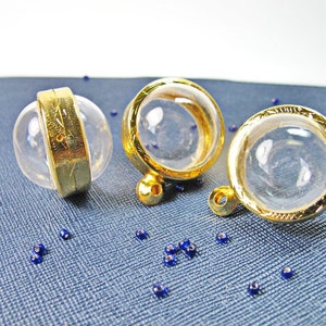 May include: Three clear glass orb pendants with gold-coloured metal accents. One orb is encircled by a gold band, while the others have gold rims and small loops. Small blue beads are scattered on the blue textured surface.