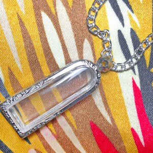 May include: A silver chain necklace with a clear glass pendant. The pendant is rectangular and has a decorative border. The necklace is lying on a patterned fabric.