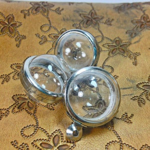May include: Three silver-toned, round, glass-top containers with clear glass lids. The containers are sitting on a brown surface with a floral pattern.