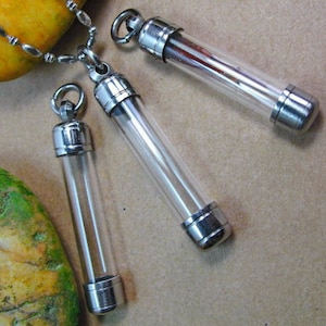 Könnte beinhalten: Drei silberne und gläserne Anhängerketten mit klaren Glasröhren und silbernen Kappen. Die Ketten liegen auf einer braunen Oberfläche.