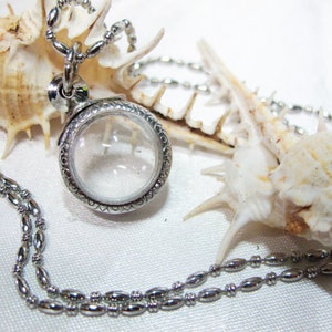 May include: A silver necklace with a round locket pendant. The locket has a clear glass window and is surrounded by a decorative border. The necklace is lying on a white surface with two seashells.
