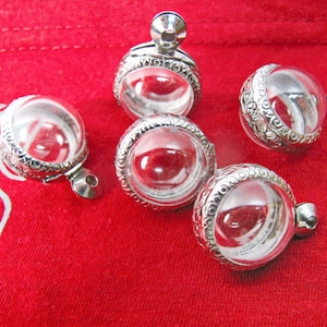 May include: Five silver and glass round pendants with intricate designs. The pendants are open and have a clear glass interior. The pendants are on a red fabric background.
