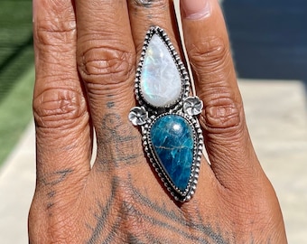 May include: A silver ring with a teardrop-shaped blue gemstone and a teardrop-shaped white gemstone. The ring is set with silver flowers.