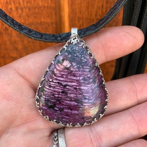May include: A teardrop-shaped pendant with a purple and black gemstone set in a silver filigree frame. The pendant is hanging from a black leather cord.