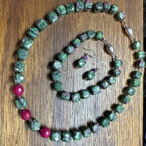May include: A green and red gemstone necklace and bracelet set with matching earrings. The necklace and bracelet are made of square-shaped beads, while the earrings are teardrop-shaped.